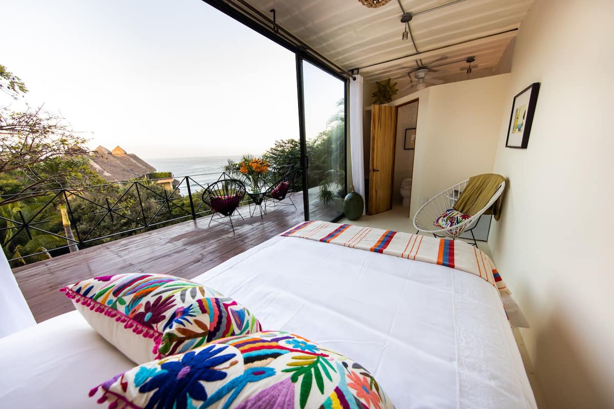 Ein Schlafzimmer mit einem großen Bett, farbenfrohen Kissen, einem Sessel und Blick auf den Ozean.