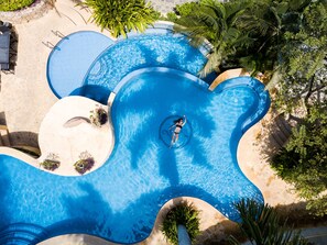 Una piscina grande y de forma original, con una persona nadando, rodeada de senderos de piedra y abundante vegetación.