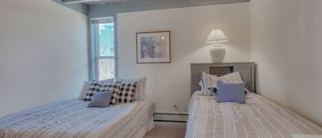 A bedroom with two beds, a window, a framed picture, and a bedside lamp.