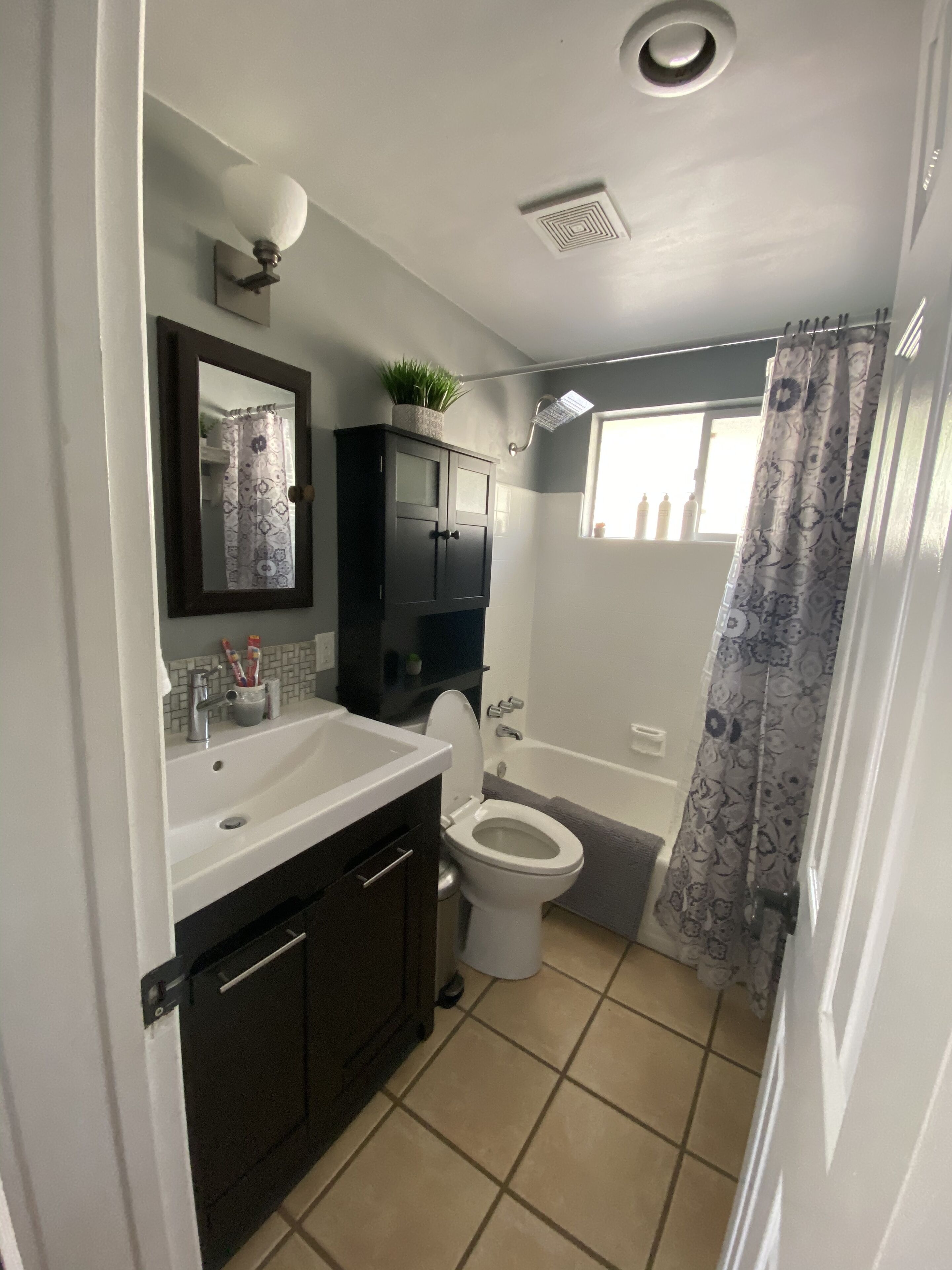 A bathroom with a white sink, dark cabinets, a toilet, a shower with a patterned curtain, and a ceiling light.
