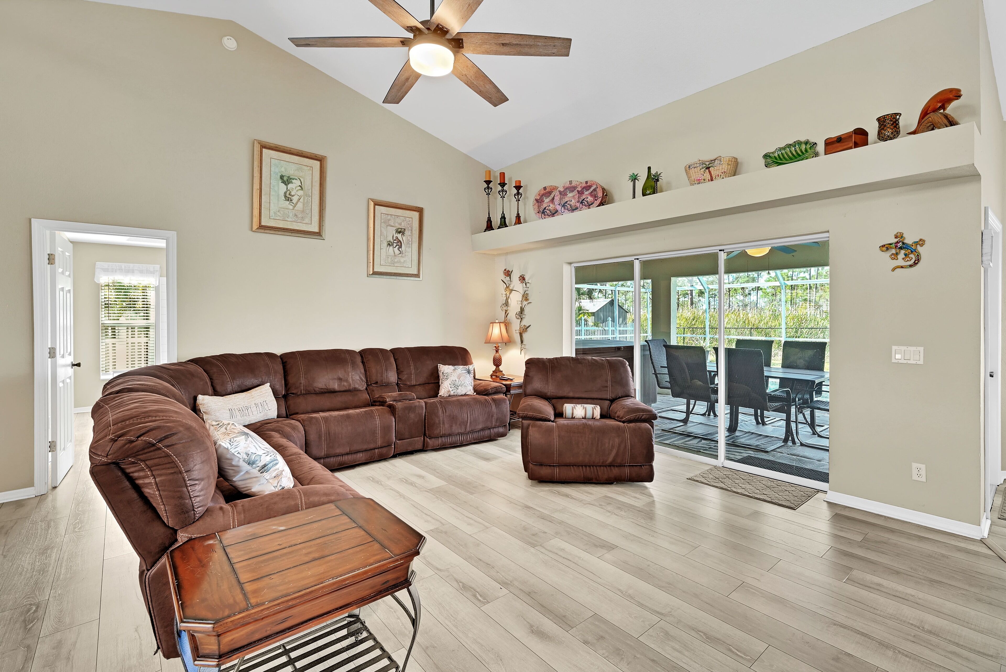 Un salon spacieux avec un canapé d’angle, un fauteuil inclinable et une table basse en bois. On y trouve également un ventilateur de plafond, une cheminée et une grande porte-fenêtre coulissante donnant sur un patio extérieur.
