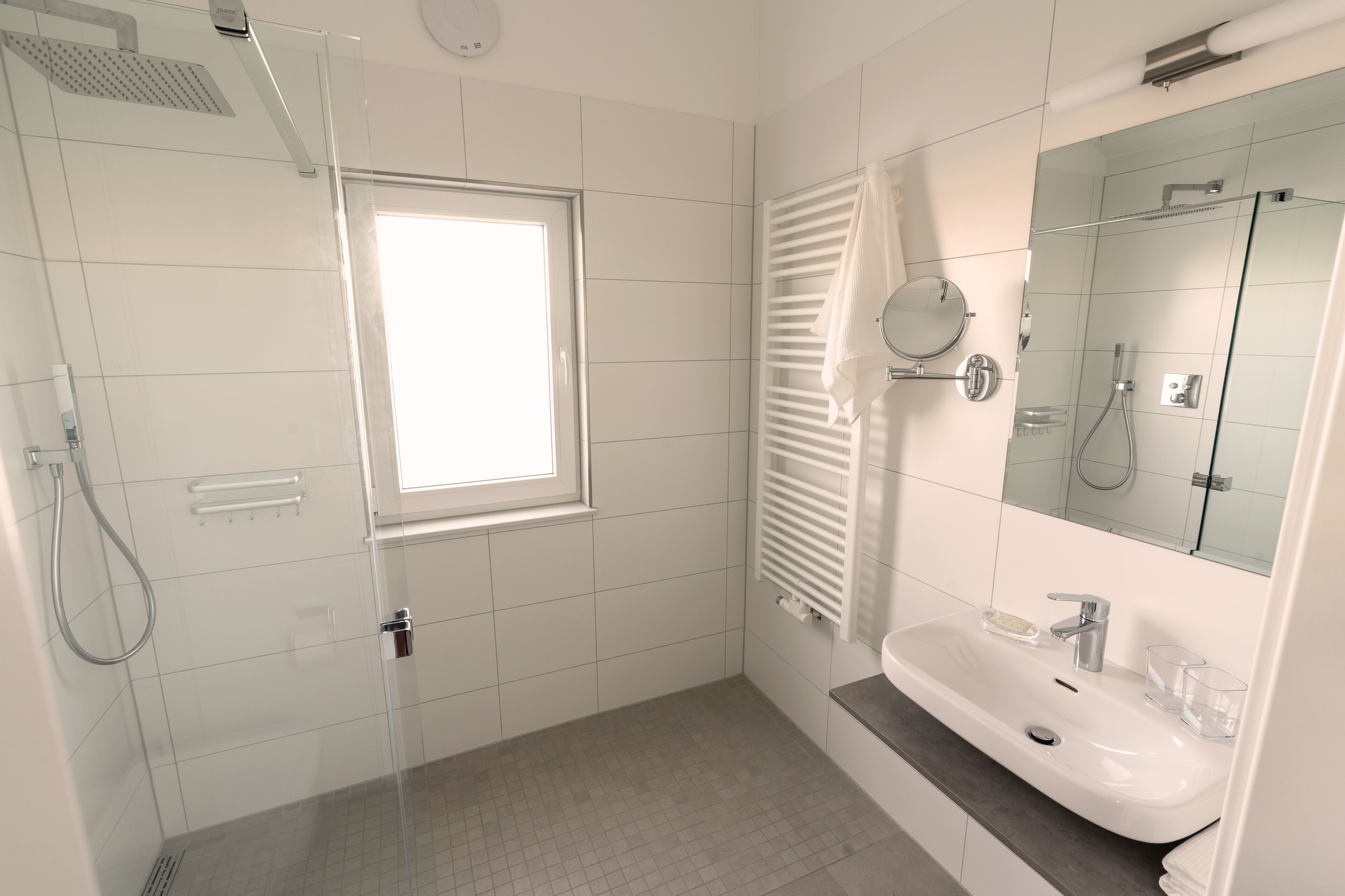 A modern bathroom with a glass shower enclosure, a window, a sink with a mirror, and a towel rack.