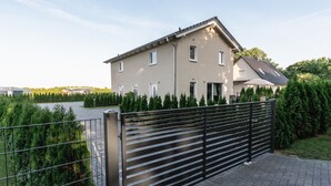 A modern two-story house with a gated driveway, surrounded by greenery and a body of water. - Ostseehaus am Naturstrand - Ostseeblick (Putgarten)