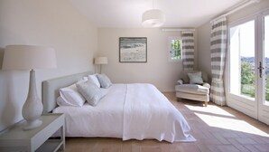 Quarto com cama, mesa de cabeceira, luminária, cadeira e uma janela grande com cortinas.