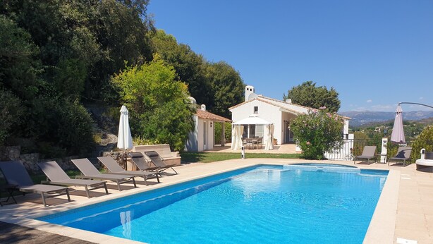 Uma piscina com espreguiçadeiras e guarda-sóis, rodeada por vegetação e um edifício branco.