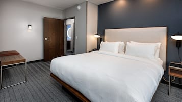 A hotel room with a large bed, a wooden bench, a desk, and a nightstand with a lamp.