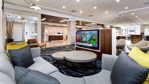 A modern hotel lobby with a seating area featuring sofas and round tables, a large flat-screen TV displaying a weather forecast, and a reception desk in the background.