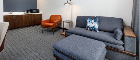 A modern hotel room with a grey sofa, a small orange armchair, a side table, a floor lamp, and a kitchenette with a microwave and coffee maker.