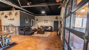 A spacious garage with a living area featuring leather sofas, a dining table, and various wall decorations. - The Dam Ranch · The Dam Ranch | Country Living + Modern Amenities (New Braunfels)