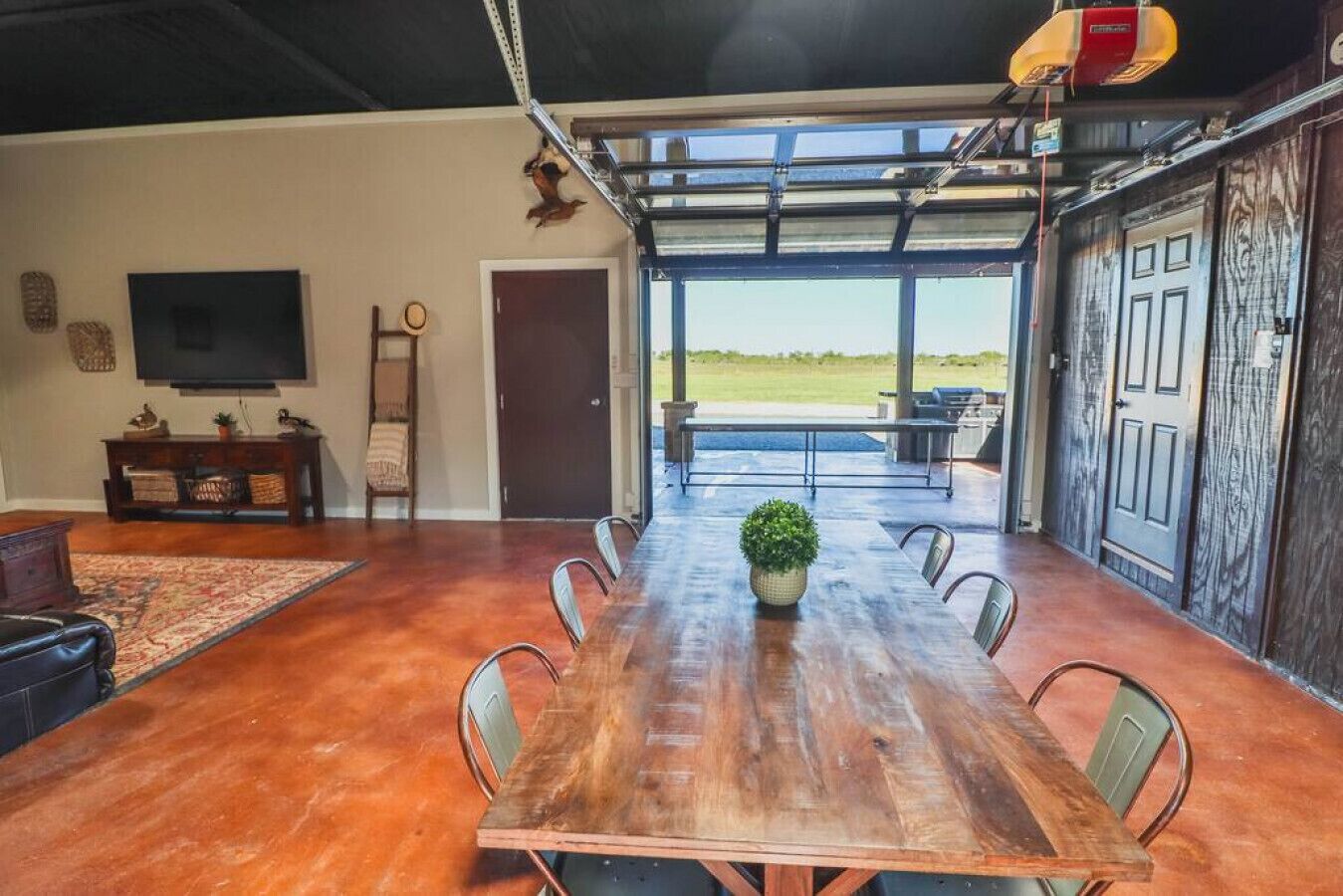 A spacious dining area with a long wooden table and chairs, a television on the wall, and a view of the outdoors through large windows.