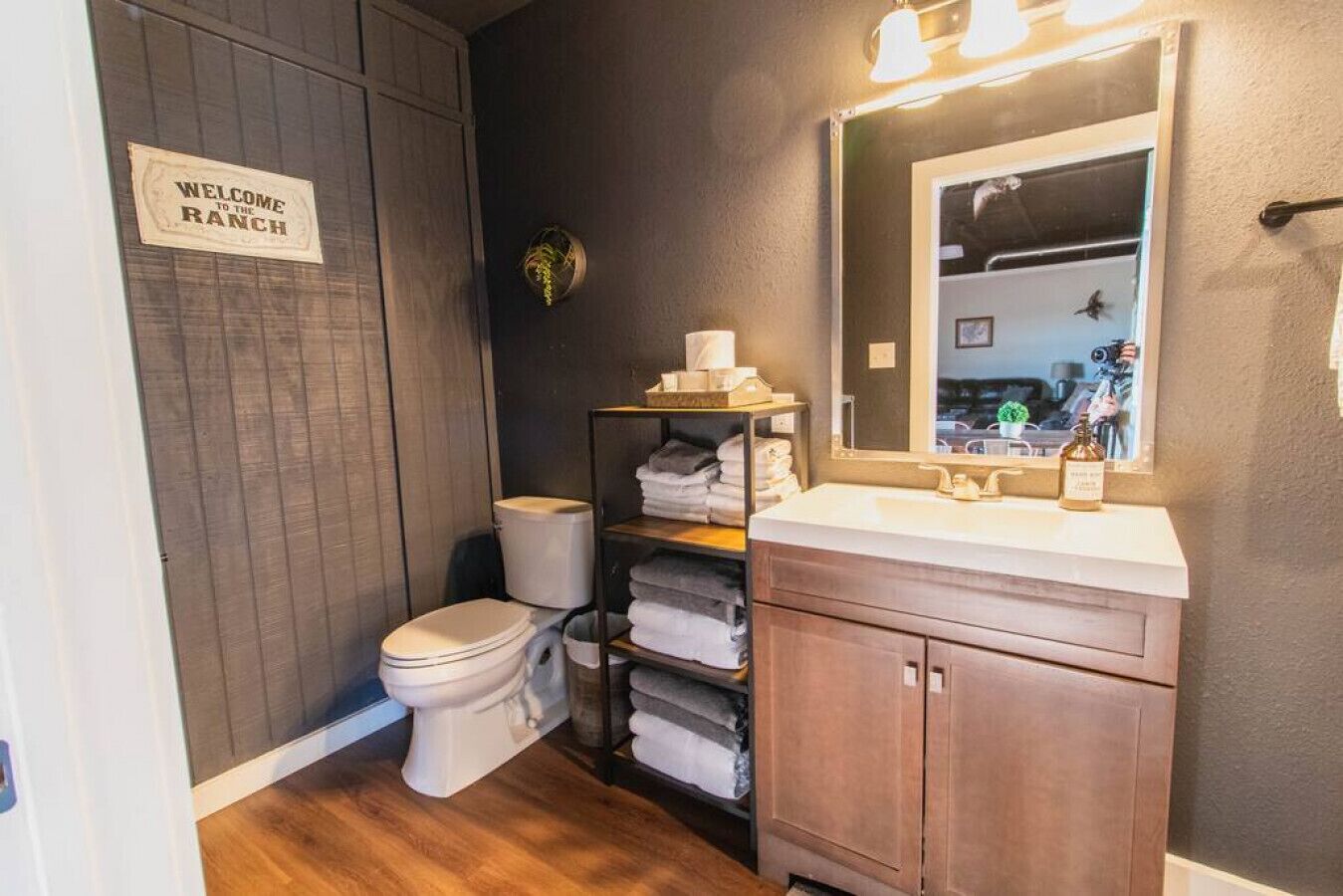 Un baño con inodoro, lavabo y estante con toallas. Hay un cartel en la pared que dice 'Welcome to the Ranch'.