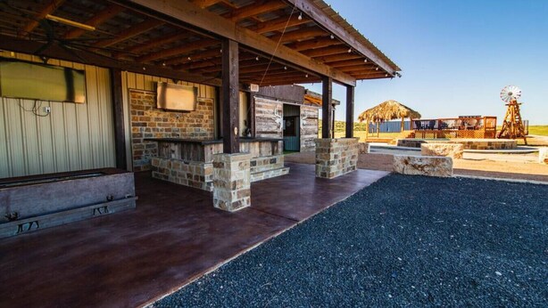 A covered outdoor area with a stone wall, a wooden deck, and a windmill in the background. - The Dam Ranch · The Dam Ranch | Country Living + Modern Amenities (New Braunfels)