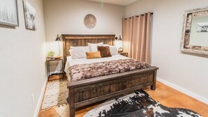 A bedroom with a wooden bed, a nightstand, a lamp, a mirror, and a cowhide rug. - The Dam Ranch · The Dam Ranch | Country Living + Modern Amenities (New Braunfels)
