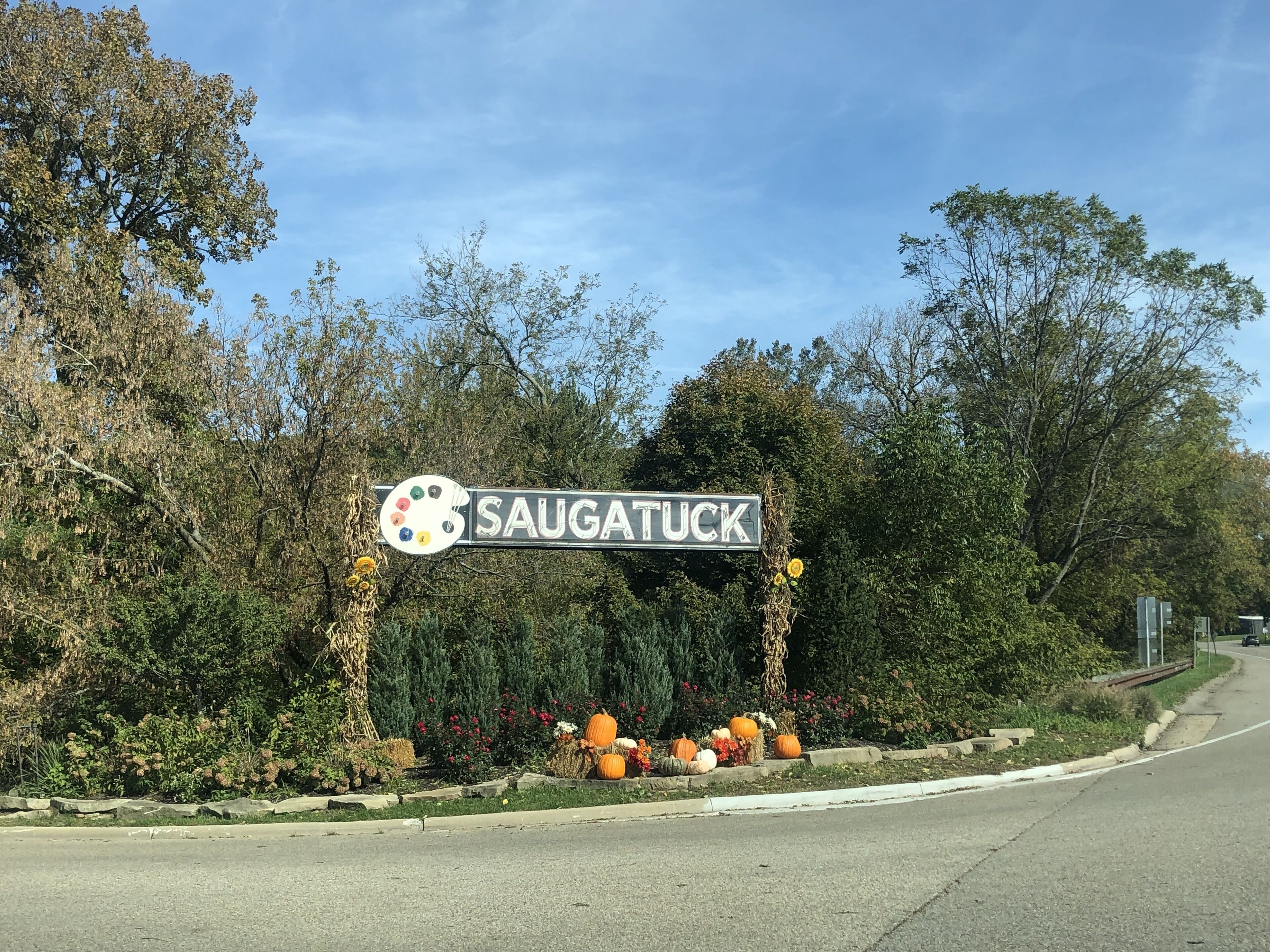 A street sign for 'SAUGATUCK' with a painter's palette logo, surrounded by autumn foliage and decorative pumpkins.