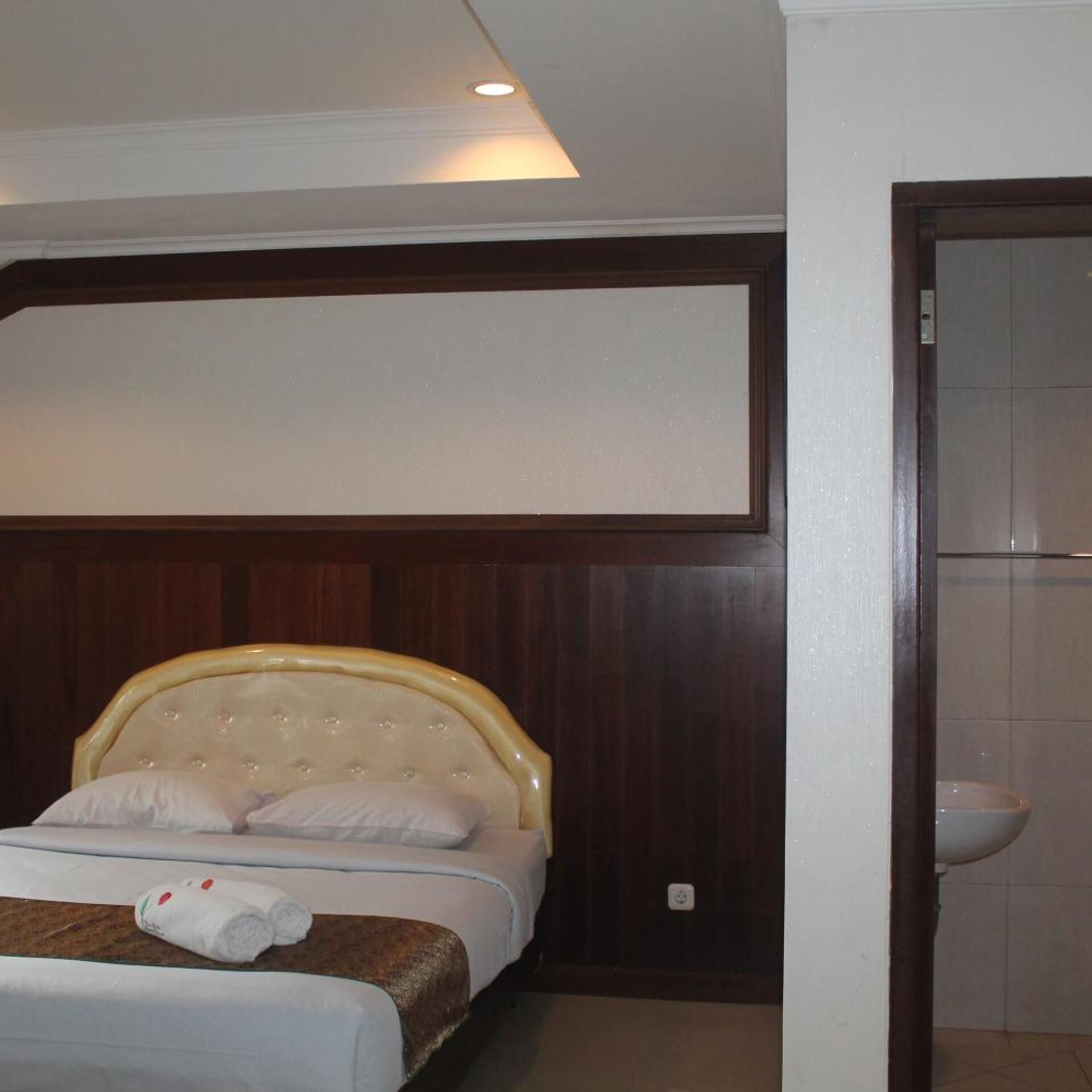 A hotel room with a bed, a headboard, a wall-mounted TV, and a bathroom with a sink and toilet.