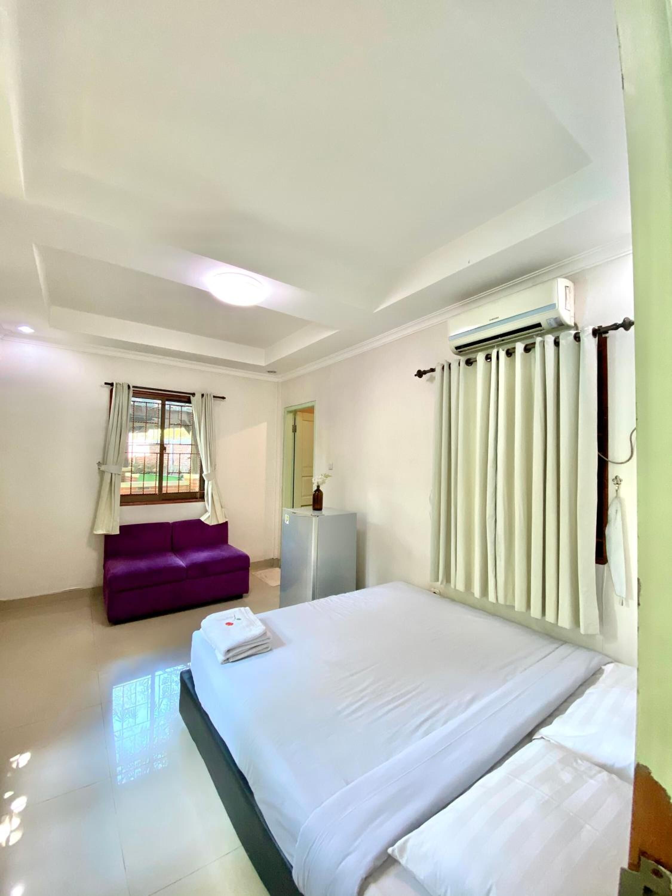 Una habitación de hotel con una cama grande, un sofá morado, un refrigerador pequeño y una ventana con cortinas.