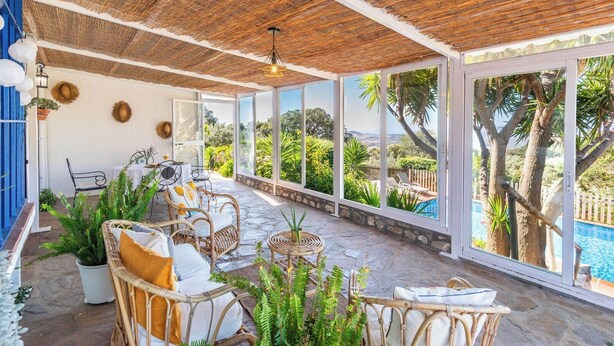 Un patio coperto con mobili in vimini, piante in vaso e vista su una piscina e alberi.