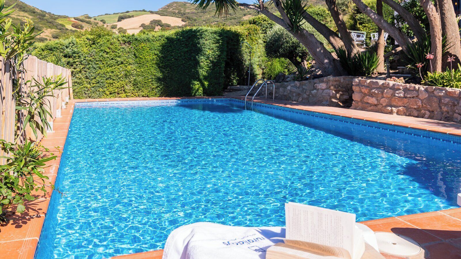 A rectangular swimming pool with clear blue water, surrounded by a stone wall and lush greenery.