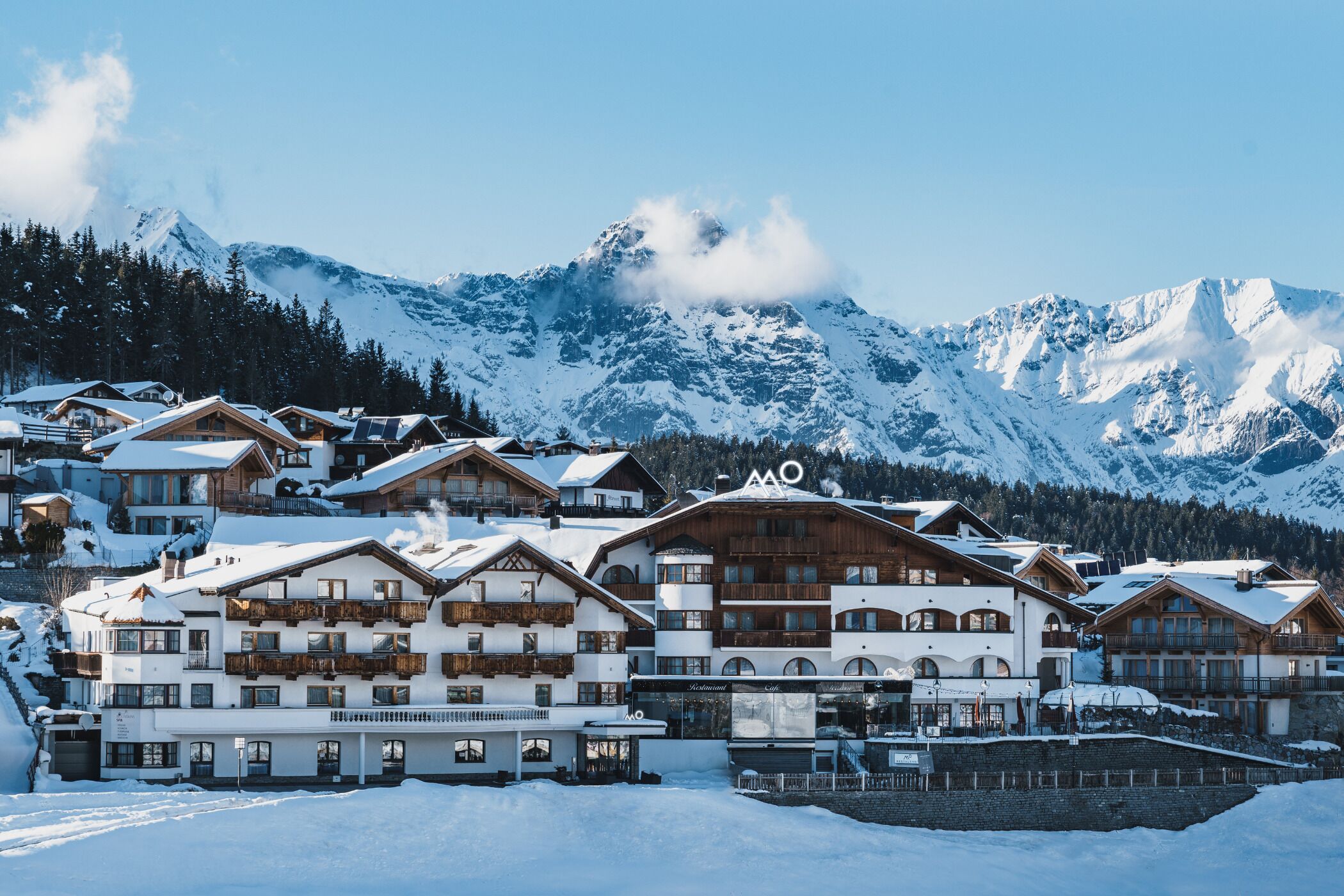 A snow-covered mountain resort with multiple chalet-style buildings, a ski lift, and a clear view of the surrounding mountains.