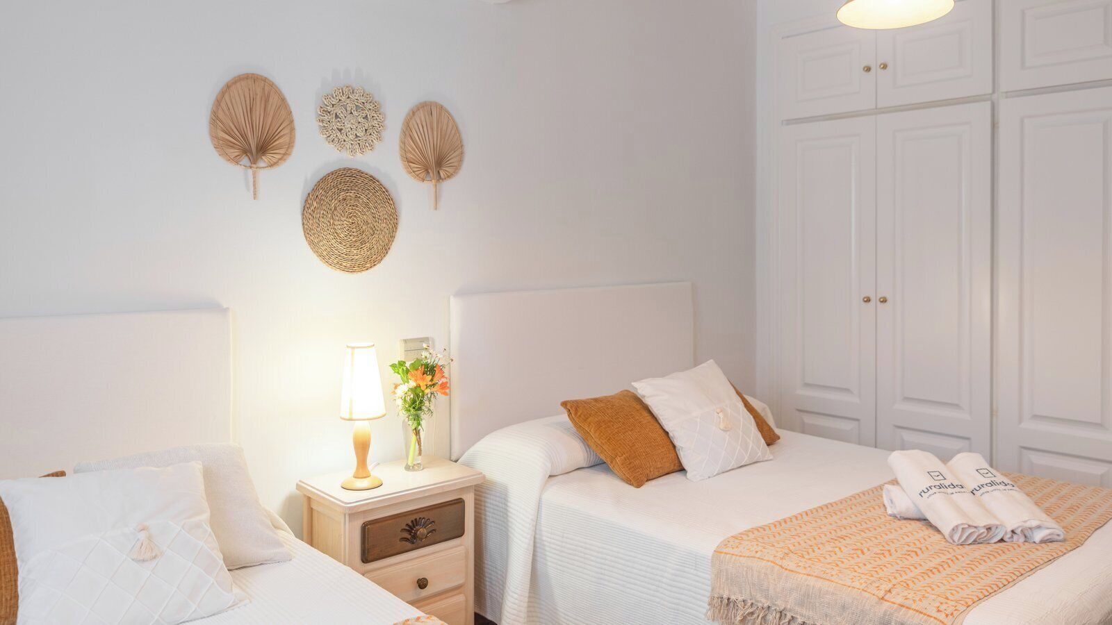 A hotel room with two beds, a nightstand with a lamp, a vase with flowers, and decorative wall hangings.
