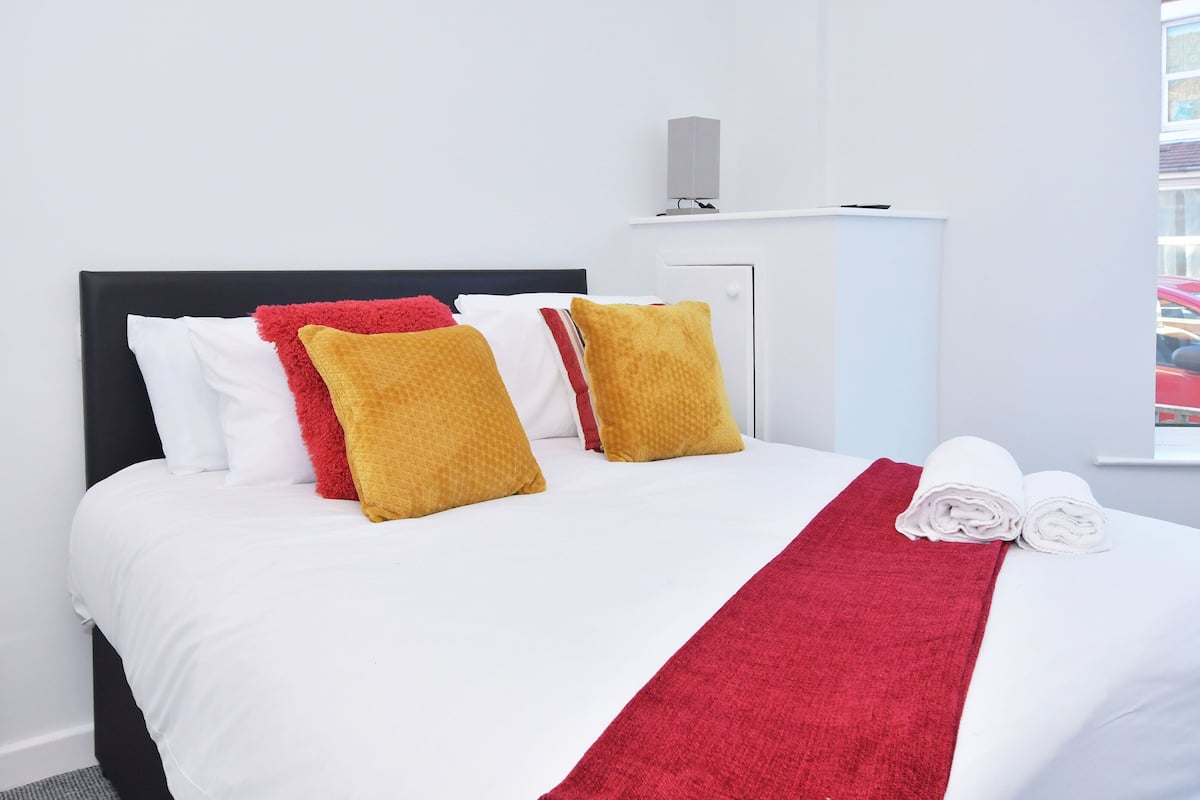 A neatly made bed with white and red bedding, a red throw blanket, and two rolled towels.