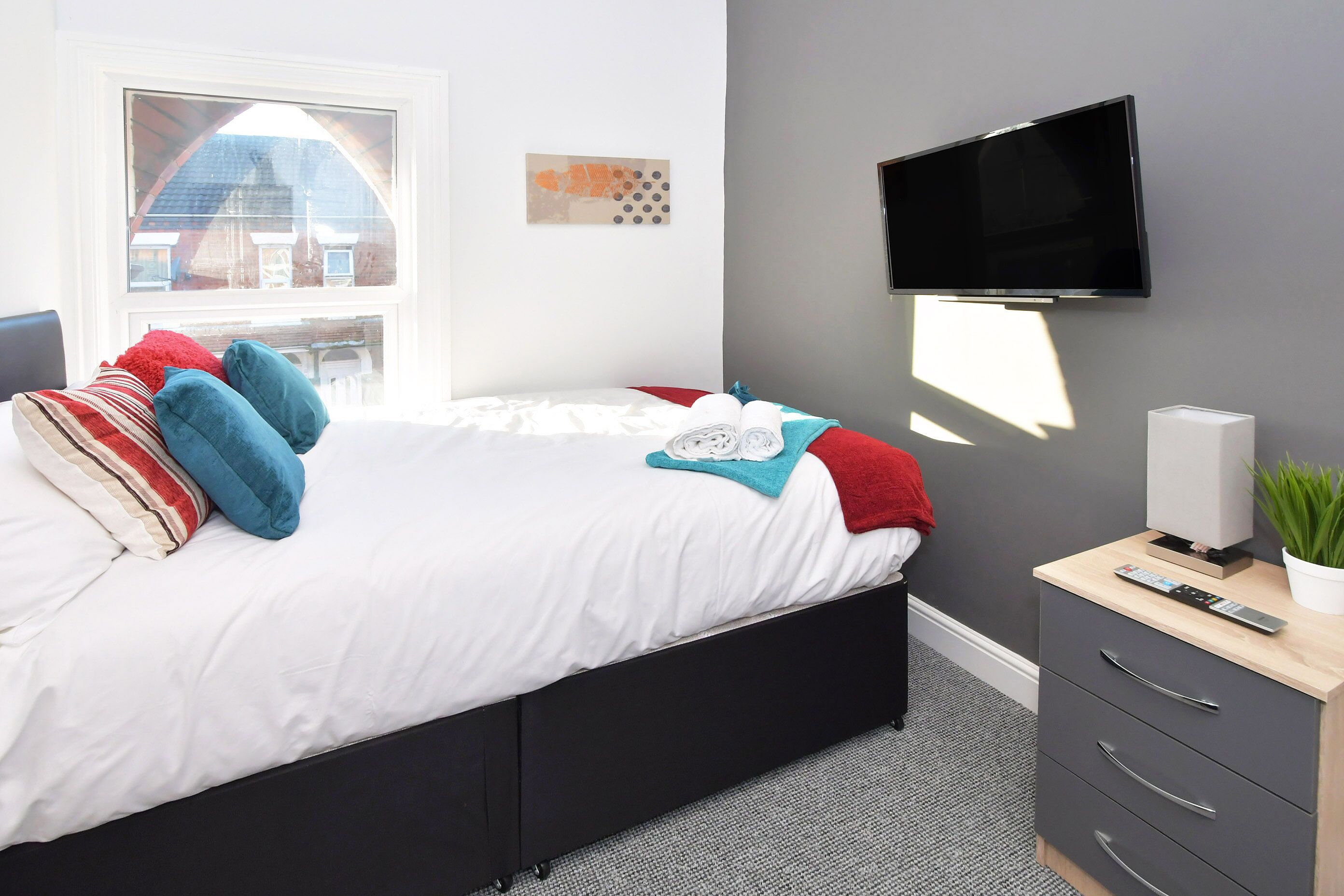 Quarto moderno com cama, televisão na parede, mesa de cabeceira com controle remoto e janela com vista para prédios.