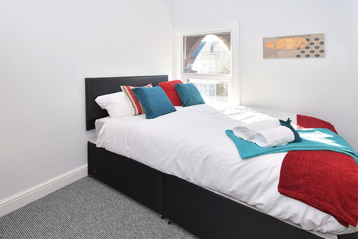 A neatly made bed with white bedding, red and blue pillows, and a red throw blanket.