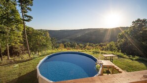 Ein Schwimmbecken mit Holzterrasse und Tisch, umgeben von üppiger Vegetation und Hügeln.