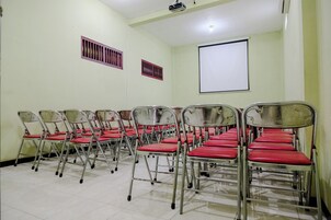 Un salón de conferencias con hileras de sillas plegables, una pantalla de proyector y rejillas de ventilación en las paredes.