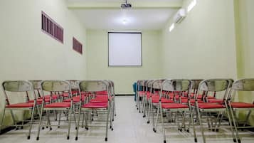Une salle de conférence avec des rangées de chaises pliantes, un écran de projection et des grilles de ventilation sur les murs.