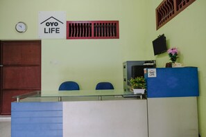 Uma área de recepção com balcão de vidro, duas cadeiras azuis, um relógio, uma ventilação, uma TV e um aviso com a inscrição "OYO LIFE".