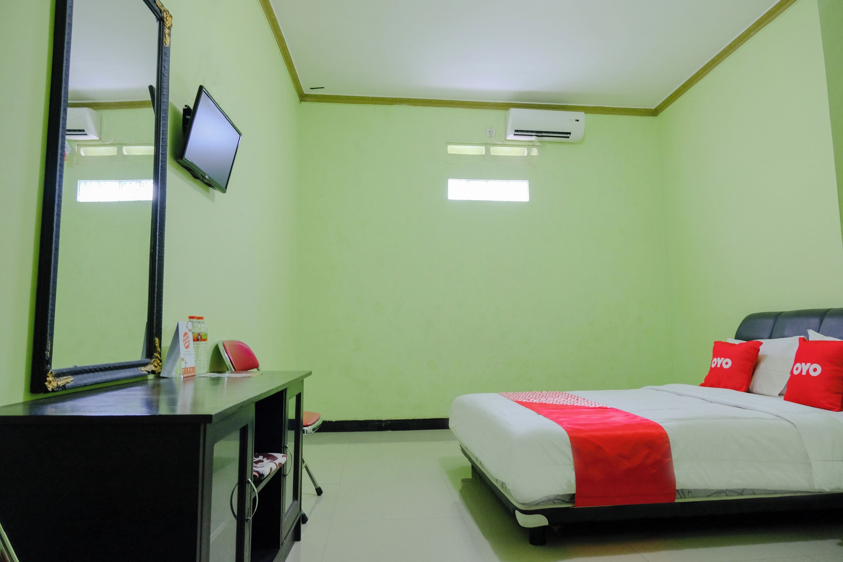 A hotel room with a bed, a desk with a mirror, a television, and an air conditioning unit.