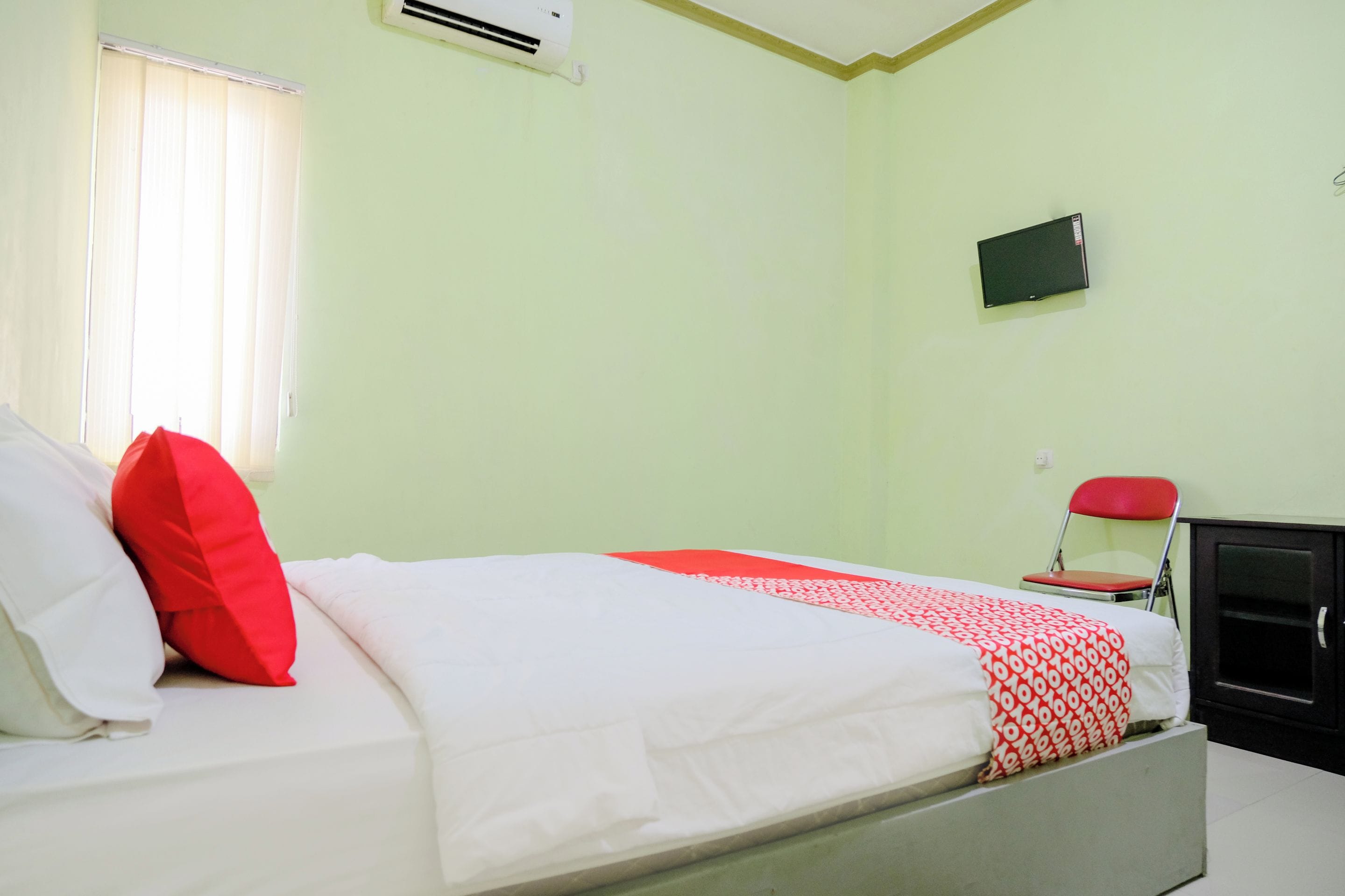 A single bed with white and red bedding, a television mounted on the wall, and a red chair.