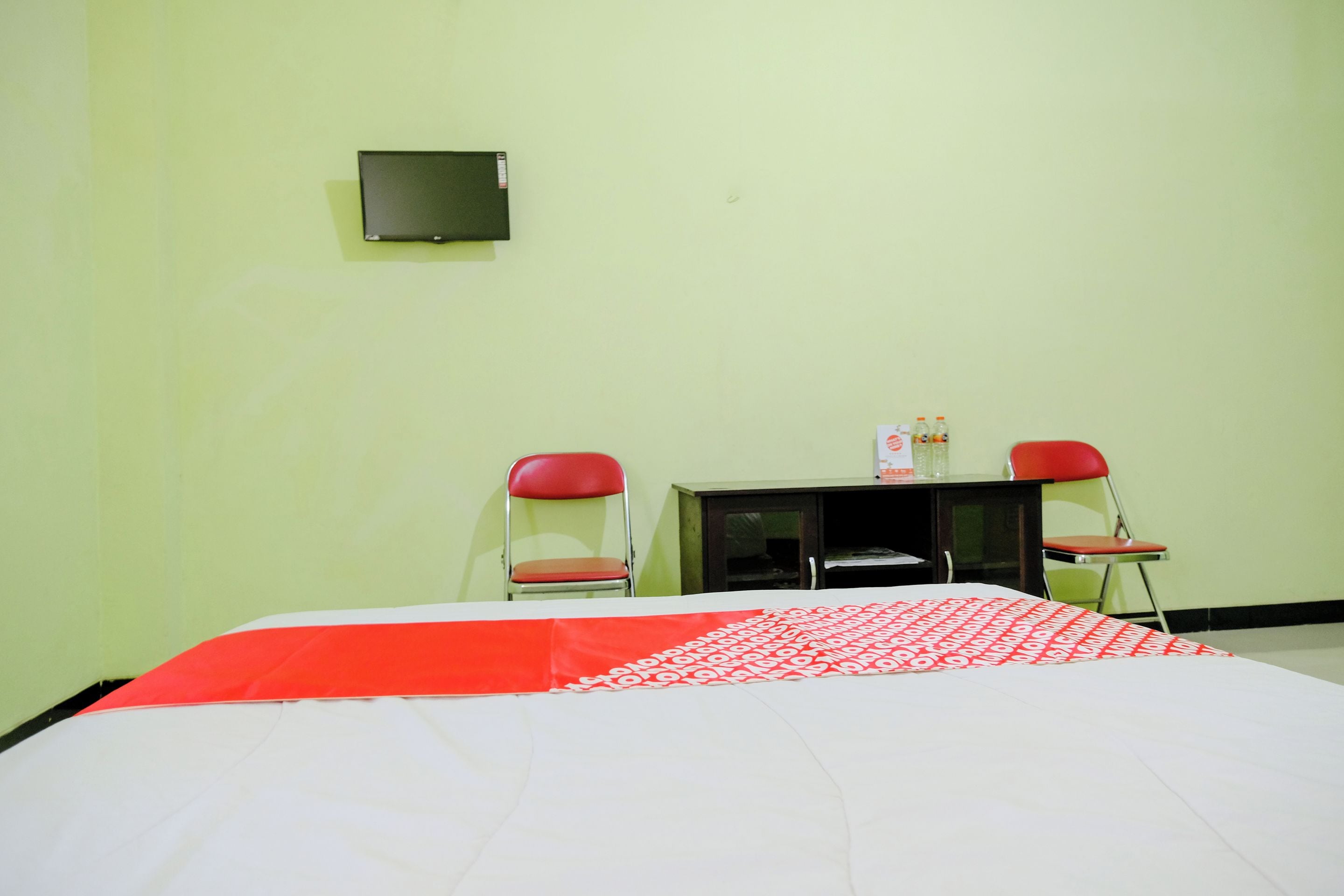 Une pièce comprenant un lit, deux chaises rouges, une petite table et un téléviseur fixé au mur.