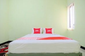 Habitación con una cama individual, una colcha blanca, dos almohadas rojas con la marca 'OYO' y una ventana con persianas.