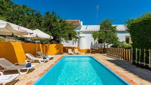 Una piscina rettangolare con lettini e ombrelloni bianchi, circondata da un muro giallo e alberi.
