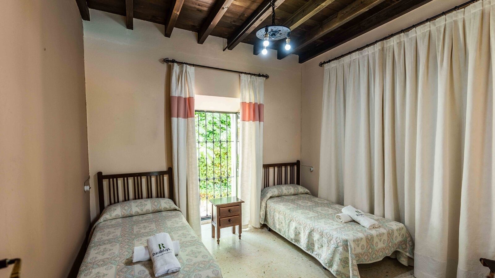 A bedroom with two single beds, a small bedside table, a window with curtains, and a wooden ceiling.