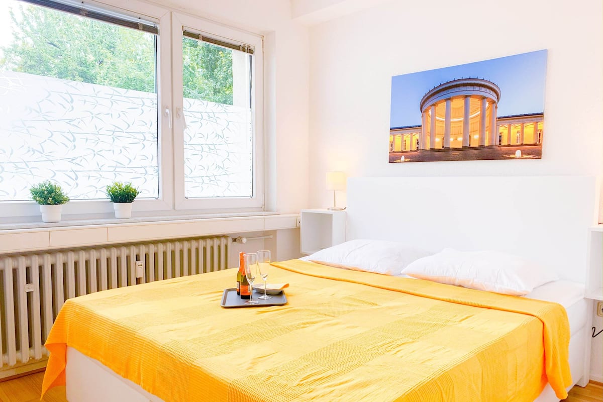 Quarto de hotel com cama, janela com persianas, radiador, mesa de cabeceira com abajur e um quadro na parede.