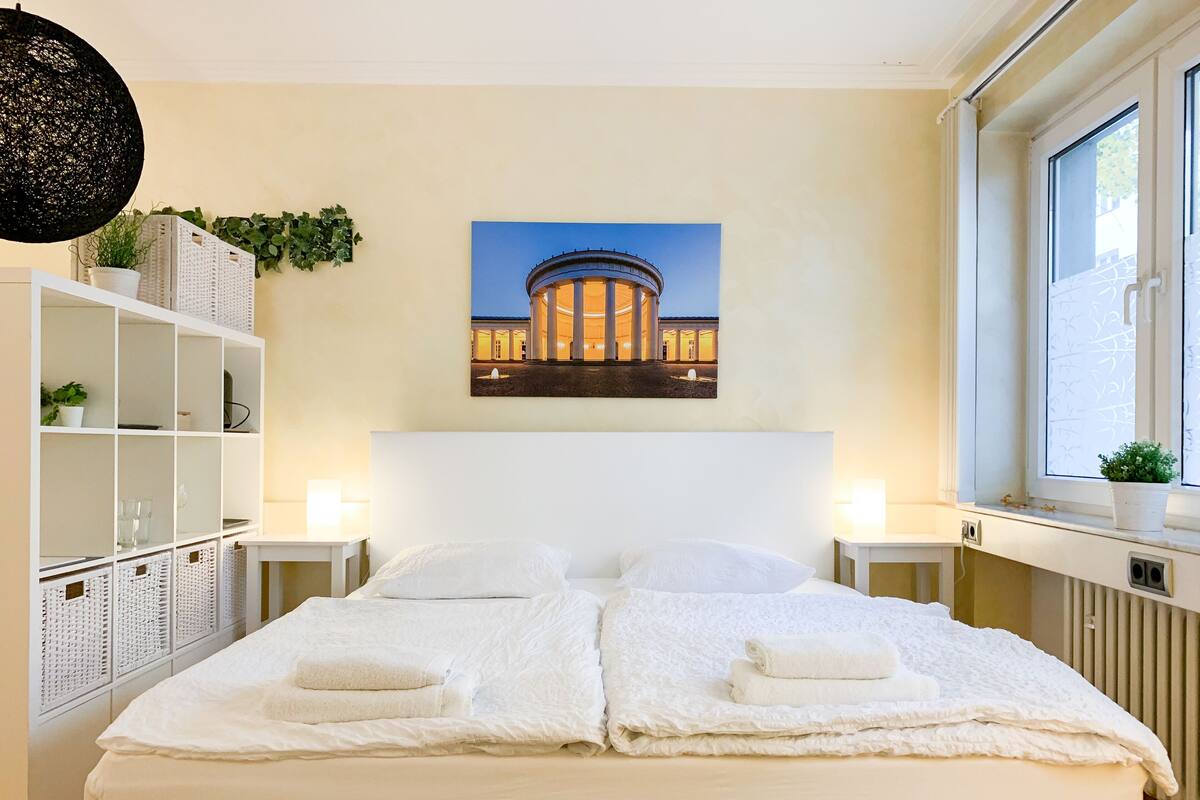 Uma cama de casal com lençóis brancos e duas almofadas, com dois mesinhas de cabeceira brancas com luminárias, uma estante branca com cestos e uma moldura com uma foto de um edifício na parede.