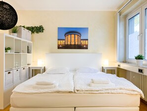 Un lit double avec du linge de lit blanc et deux oreillers, de chaque côté duquel se trouvent des tables de chevet blanches avec des lampes, une étagère blanche avec des paniers, et un tableau représentant un bâtiment accroché au mur.