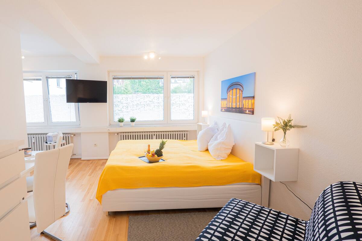 Quarto de hotel com cama, televisão, janela com plantas e um quadro na parede.
