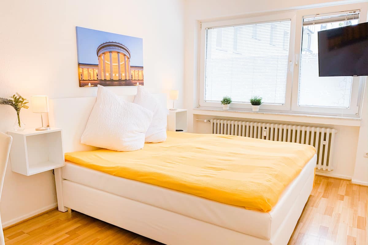 Quarto com cama, mesa de cabeceira, janela com persianas, radiador e televisão na parede.