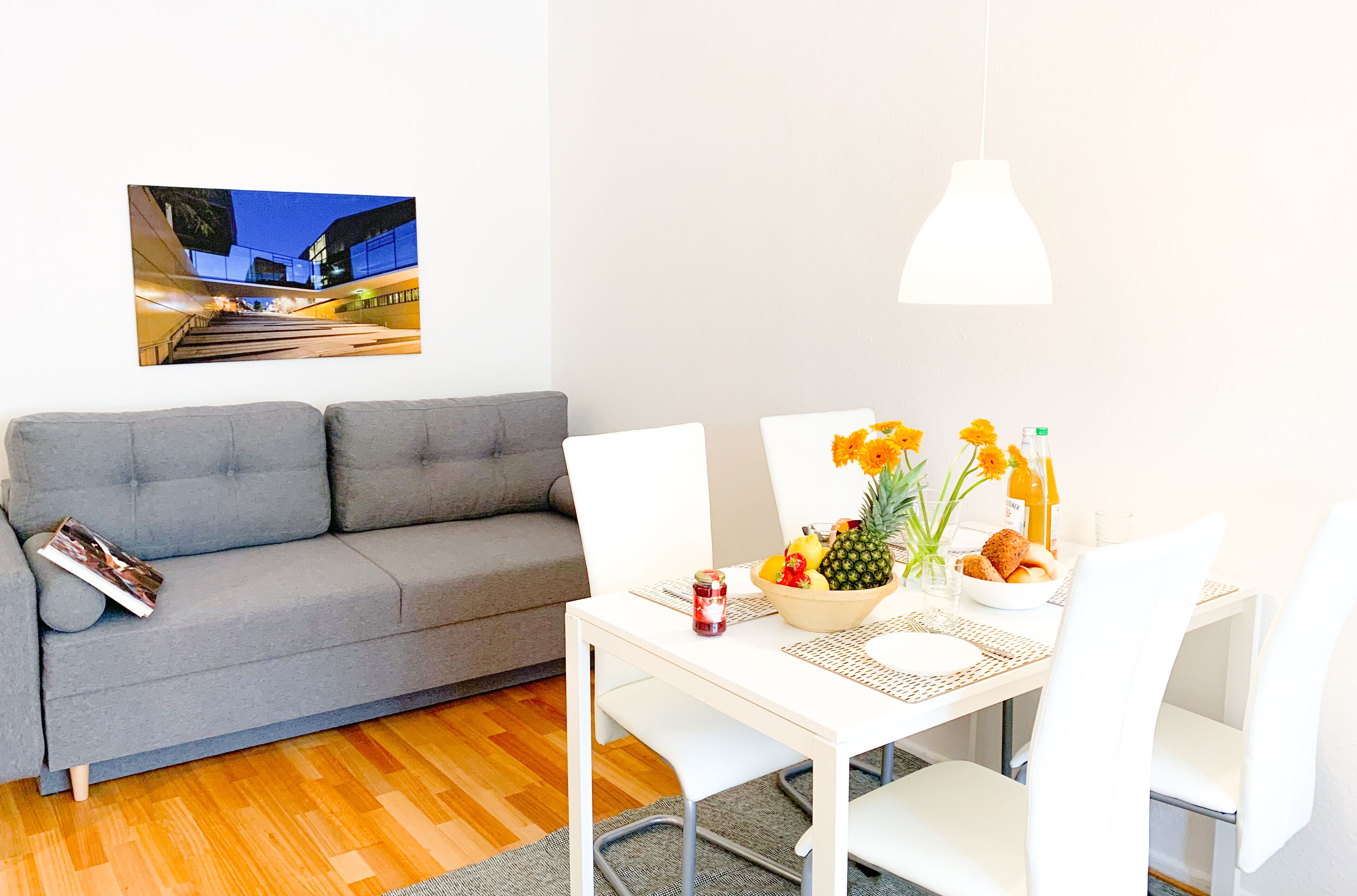 Una sala de estar moderna con un sofá gris, una mesa de comedor con sillas blancas, un frutero y una lámpara colgante.
