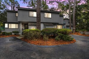 A two-story modern house with a driveway, surrounded by trees and shrubs.