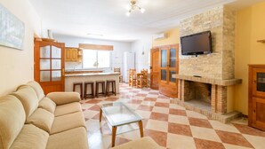 A living room with a sofa, a glass coffee table, a checkered floor, a kitchen area with a stone fireplace, and a TV mounted on the wall.