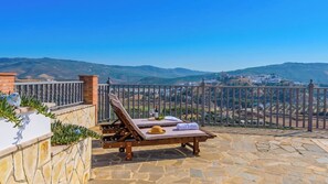 A terrace with lounge chairs and a table, overlooking a scenic landscape with hills and a distant town. - El Mirador de Iznájar by Ruralidays (Córdoba)