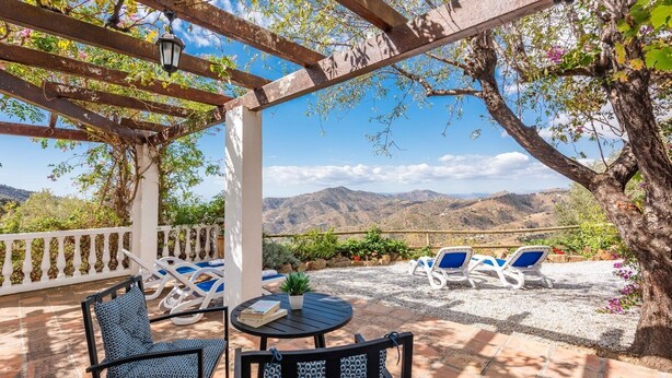 Un espacio de estar techado al aire libre con mesas y sillas, con vistas a un paisaje montañoso.