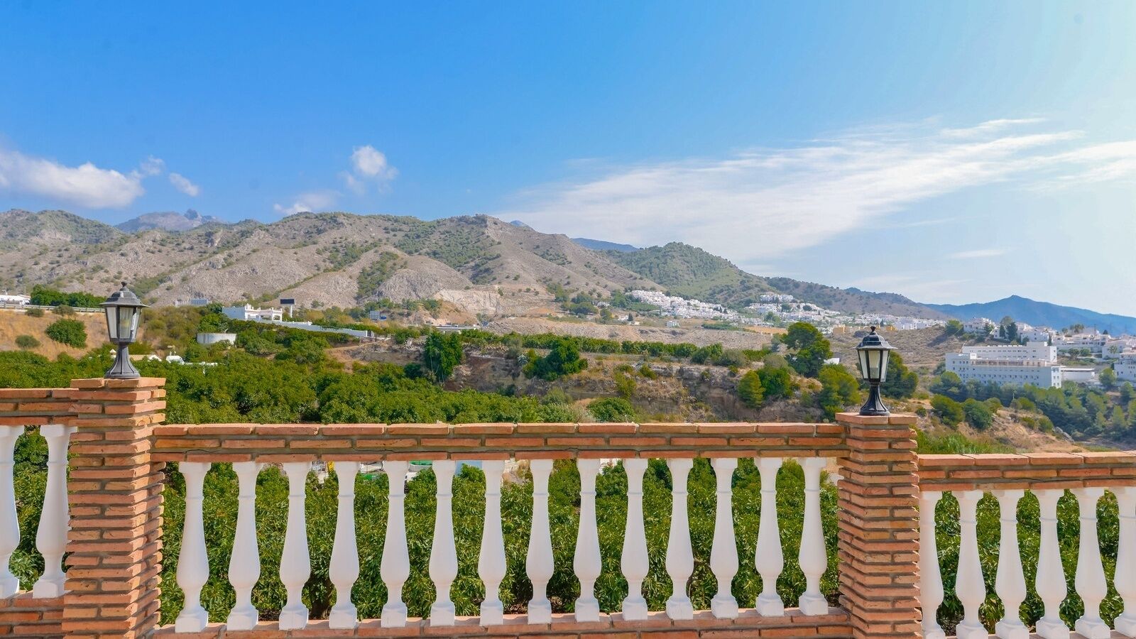 A balcony with a brick railing and white balusters, overlooking a mountainous landscape with greenery and buildings in the distance.