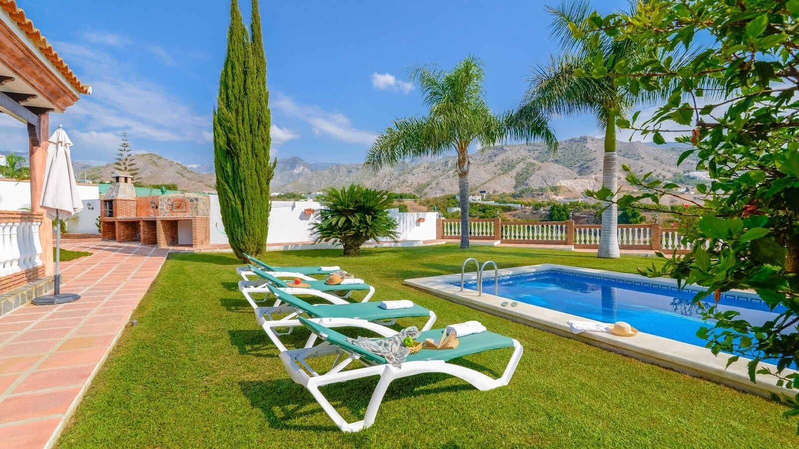 A pool area with lounge chairs, a barbecue, and a view of hills.