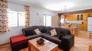A living room with a sofa, a coffee table, and a red ottoman. There is a kitchen area with wooden cabinets and a dining table. - Villa Pastora Nerja by Ruralidays (Málaga)